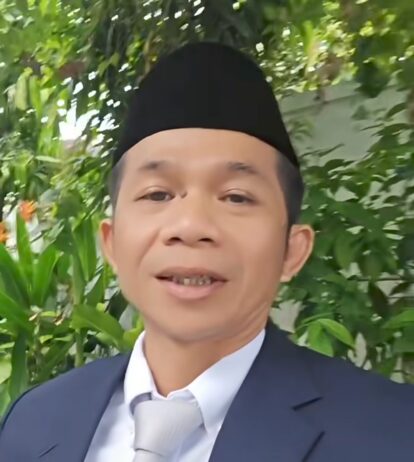 Smiling man in a dark suit and light tie, wearing a black cap, outdoors with green plants in the background.